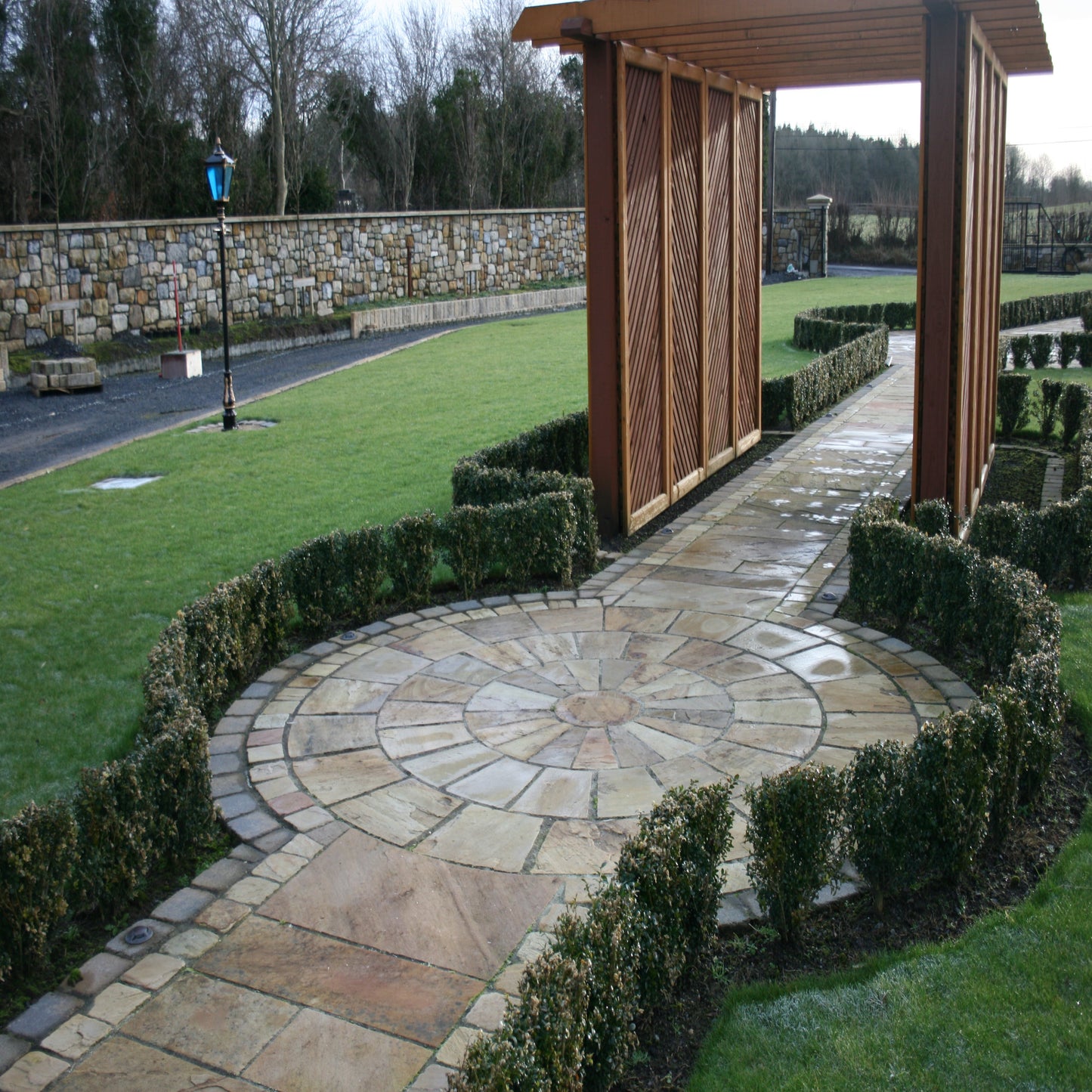 A circular pathway design completed using Tinted Mint Sandstone Paving Circle with a wooden structure and manicured hedges in the background.