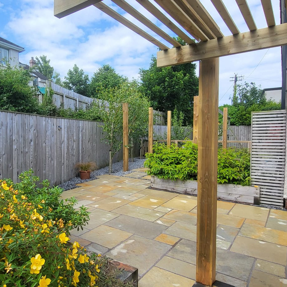 Backyard with stone patio, wooden pergola, and greenery under a blue sky. Paving installed is Yellow Limestone Paving Tumbled Patio Packs. 