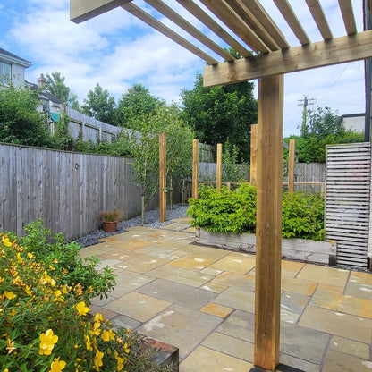 Backyard with stone patio, wooden pergola, and greenery under a blue sky. Paving installed is Yellow Limestone Paving Tumbled Patio Packs. 
