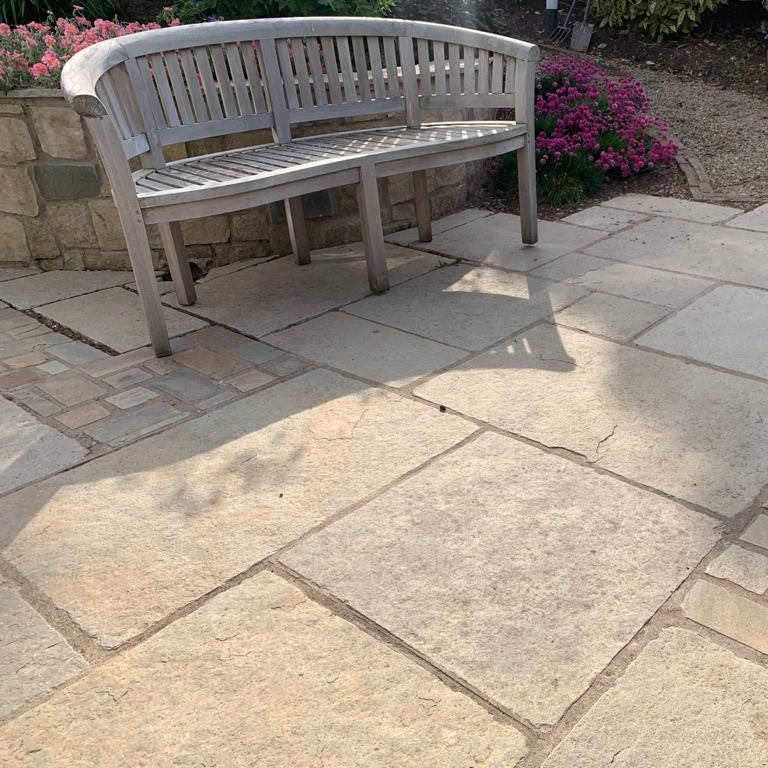 Wooden bench on a stone patio with pink flowers in the background. Paving is tumbled Yellow Limestone. 