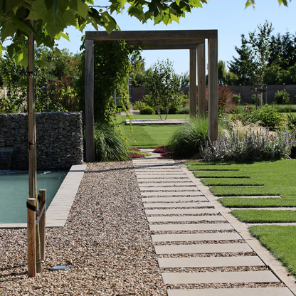 A modern outdoor garden scene with a gravel path and stone slab walkway leading toward a wooden arched pergola over a landscaped lawn, with a pool edge visible on the left and leafy branches framing the top of the image.