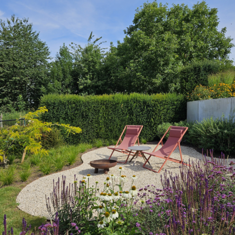 A landscaped garden seating area with two pink folding deck chairs on a light gravel patio, surrounded by purple flowers, green shrubs, and trees under a blue sky.